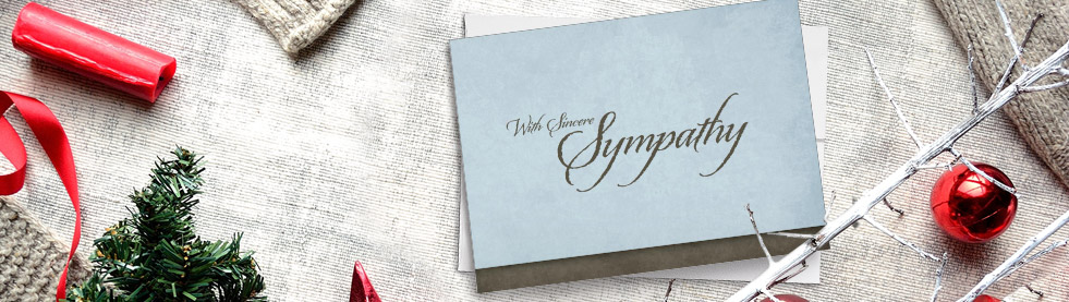 Blue sympathy card lain among Christmas objects like red ribbon, ornaments, and foliage.