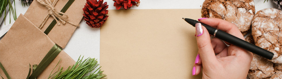 Long image of a female hand starting to write on a brown Christmas card with seasonal accents like pine sprigs, pine cones., cookies, and packages.