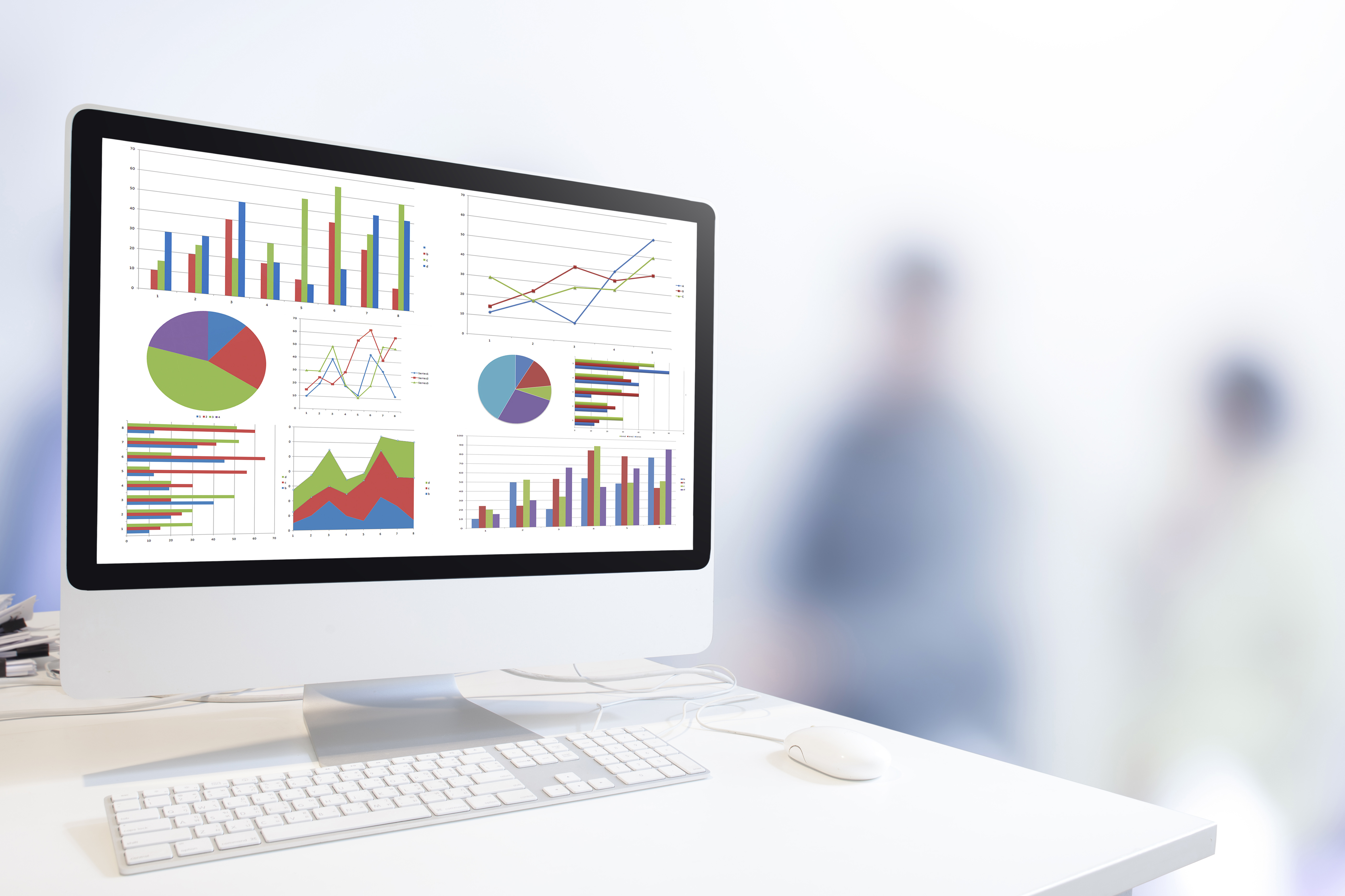 Modern desktop computer showing various multicolored graphs, with out of focus human figures in the background.