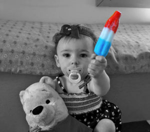 Black and white photo of a baby girl in a dress, sucking on a pacifier, holding a teddy bear in one hand and a bright red, white, and blue popsicle in the other.