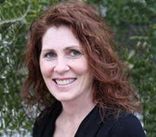 Smiling woman with auburn hair and gray eyes, with a green tree in the background.