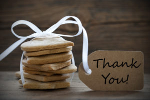 A stack of delicious light brown square-shaped cookies tied with a white ribbon and complimented with a light brown gift tag that reads 'thank you' in black letters, all against a woodgrain background.