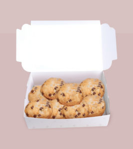 A delicious batch of chocolate chip cookies with coconut on top, in a white bakery box, against a soft pink background.