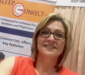Selfie of a middle-aged blonde woman in glasses and a coral-colored shirt, standing in front of a curtain and a billboard advertising for a company called Sleep Connect, Inc.