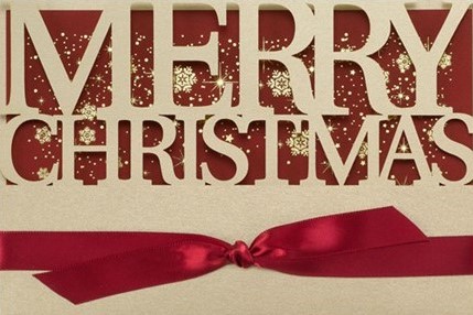A close-up shot of a beautiful holiday greeting card with a large red bow tied around the bottom. The top part of the card is laser-cut with the words 'Merry Christmas' in a muted gold color against a red background doted with golden confetti. 