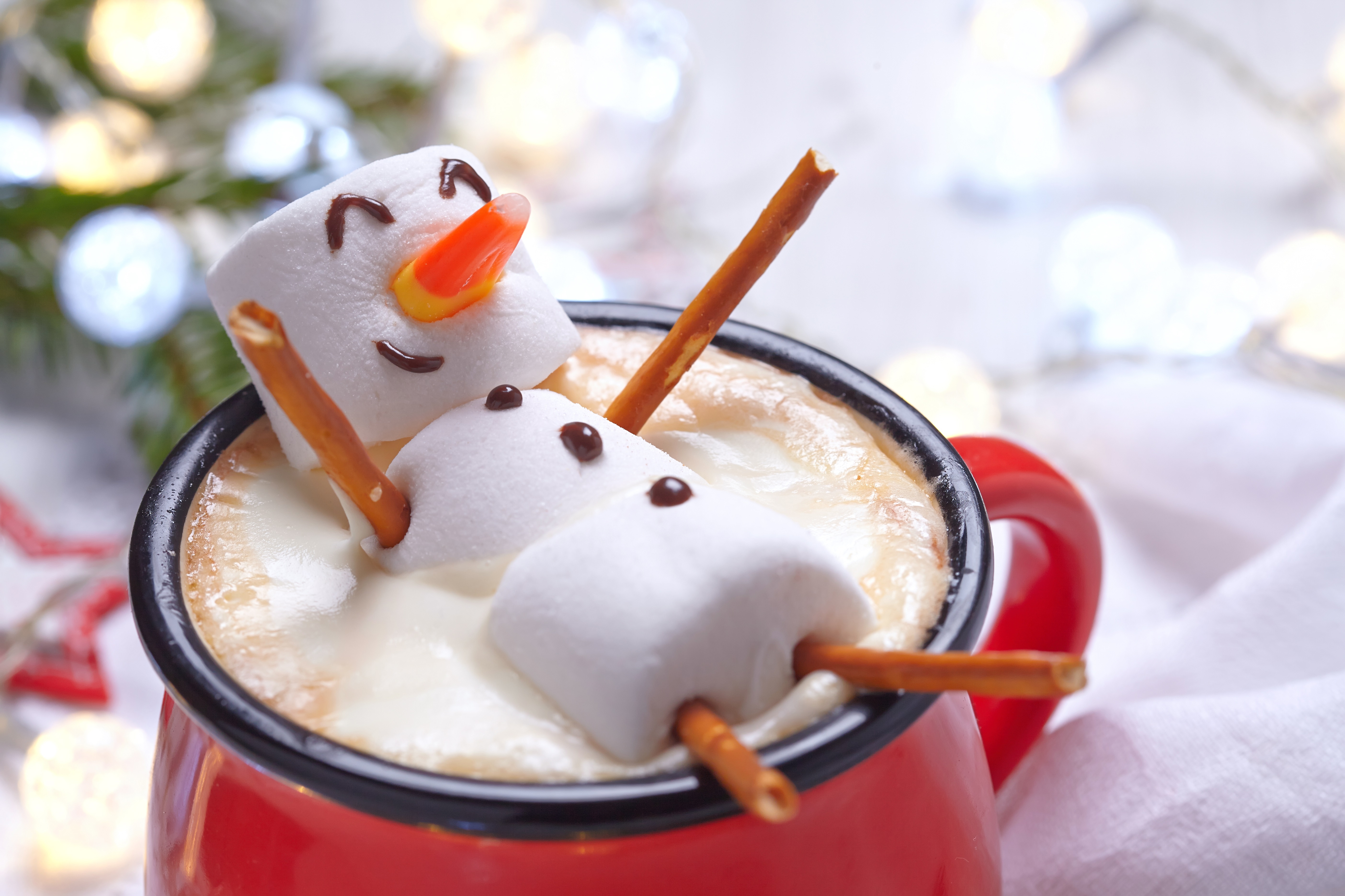 A marshmallow snowman with chocolate eyes, mouth, and buttons, a candy corn nose, and pretzel arms and legs lays in a red mug of hot chocolate at the foreground of an out-of-focus holiday background with lights, a tree, and other seasonal objects.