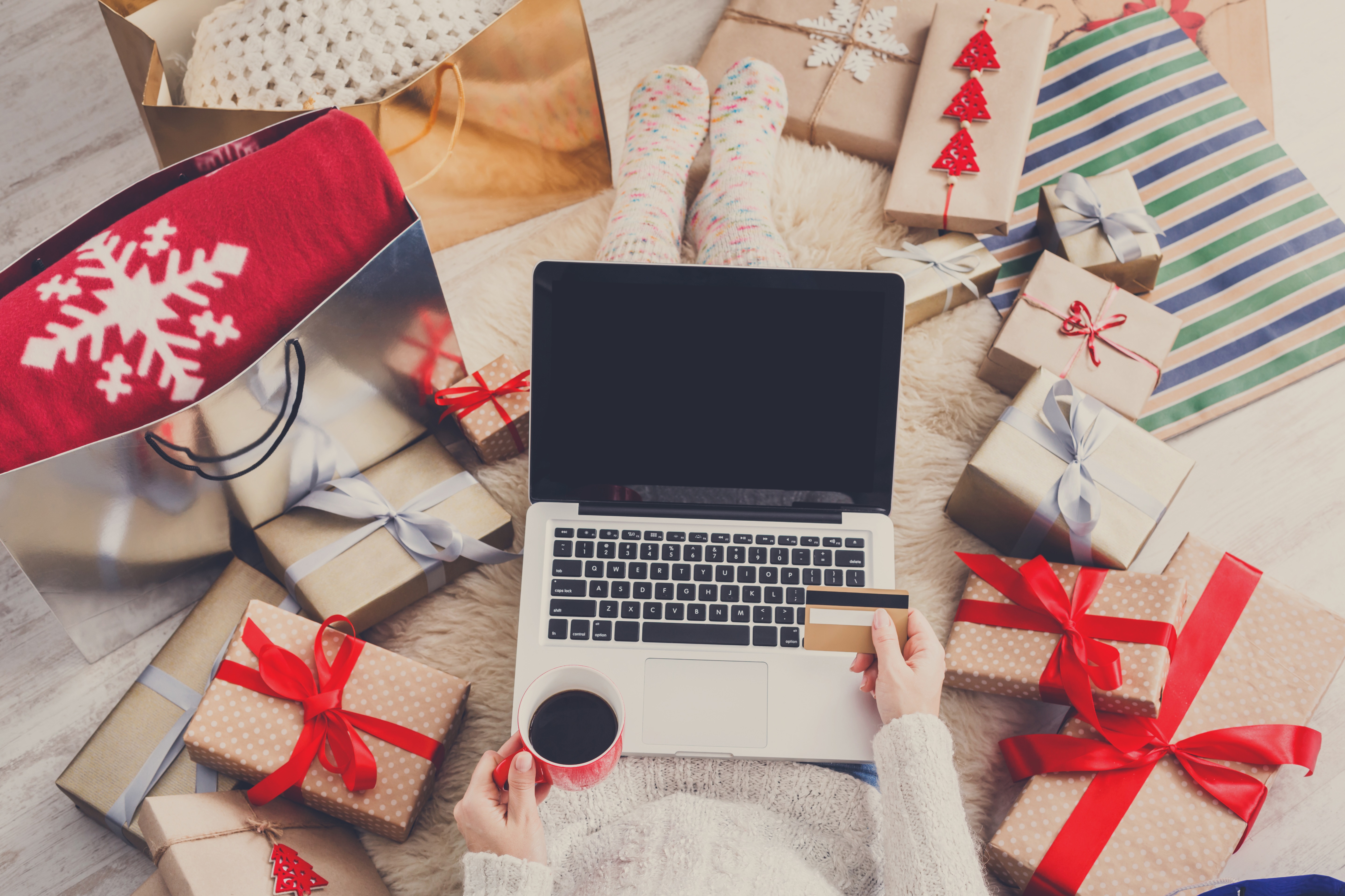 A laptop, about to be used for online shopping, is being operated from a female perspective, as she looks at her credit card and sips a cup of coffee amidst a pile of colorful Christmas gifts.