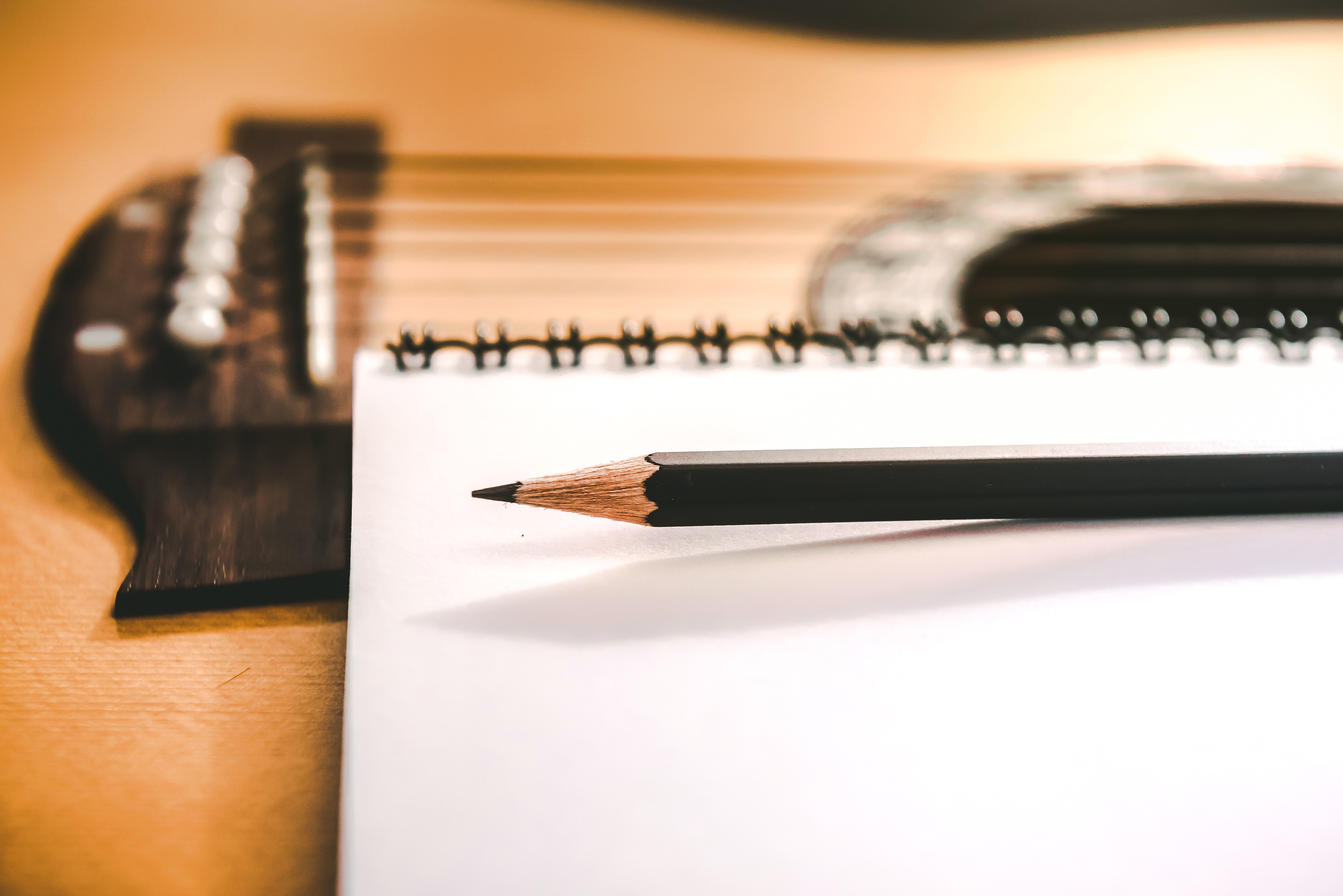 Close-up of a clean white notebook with a freshly sharpened black pencil siting on top of an acoustic guitar.