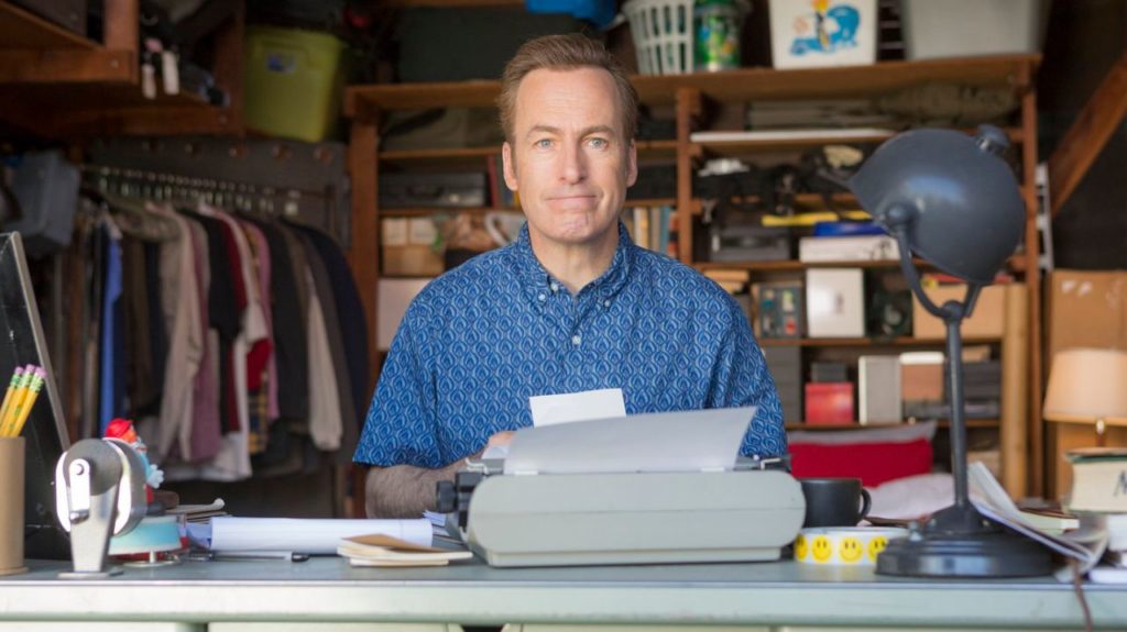 Bob Odenkirk sitting at a table in the garage, in a blue shirt, typing on a typewriter.