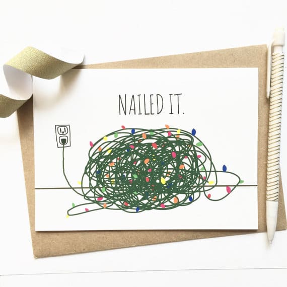 A hand-drawn greeting card featuring a pile of tangled Christmas lights with the words 'Nailed It' above it. The card lays against a brown envelope, and is accented with a bit of ribbon and a decorative pen.