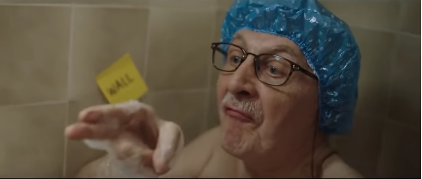 Elderly man sitting in a bathtub with a blue shower cap on.