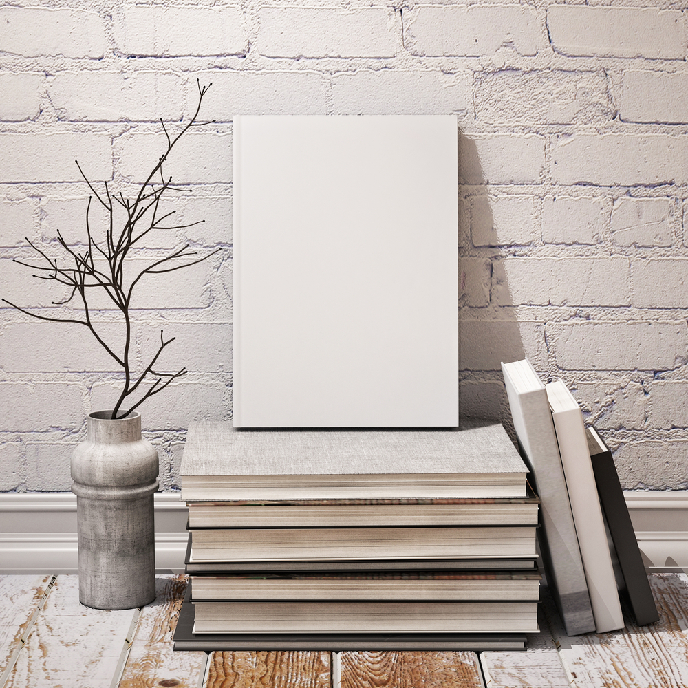 A shabby chic scene shows books in black, gray, and white tones, as well as some twigs inside a vase in similar hues, sitting on wooden floor that is partially painted white and leaning against a white brick background.