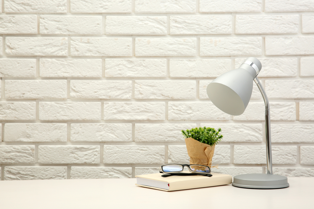 A light-silver lamp, a beige book, black-rimmed glasses, and a green plant sit on a light-colored desk against a white brick background.