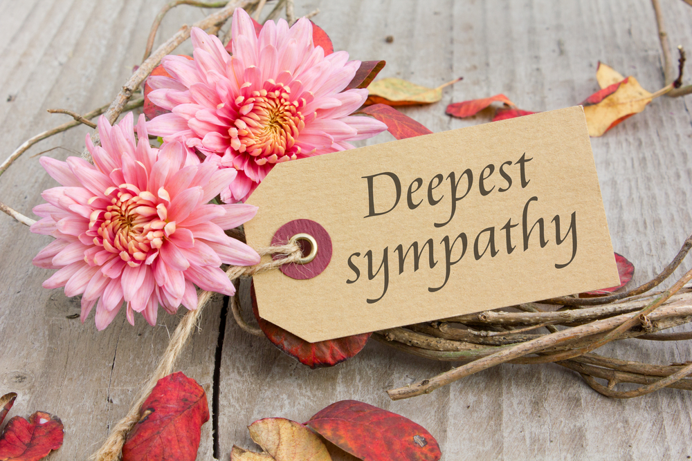 Attached to a bouquet of pink flowers and twigs with twine is a small brown tag that reads 'Deepest sympathy.' Dried red and brown leaves are scattered about a wood-grain background.