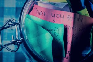 A mason jar, resting on a gingham table cloth, open with thank-you messages inside. The messages are written on different colored pieces of paper. 