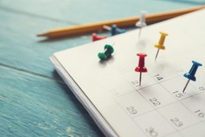 Close up of calendar on a table (next to a pencil), with colored tacks in certain days.