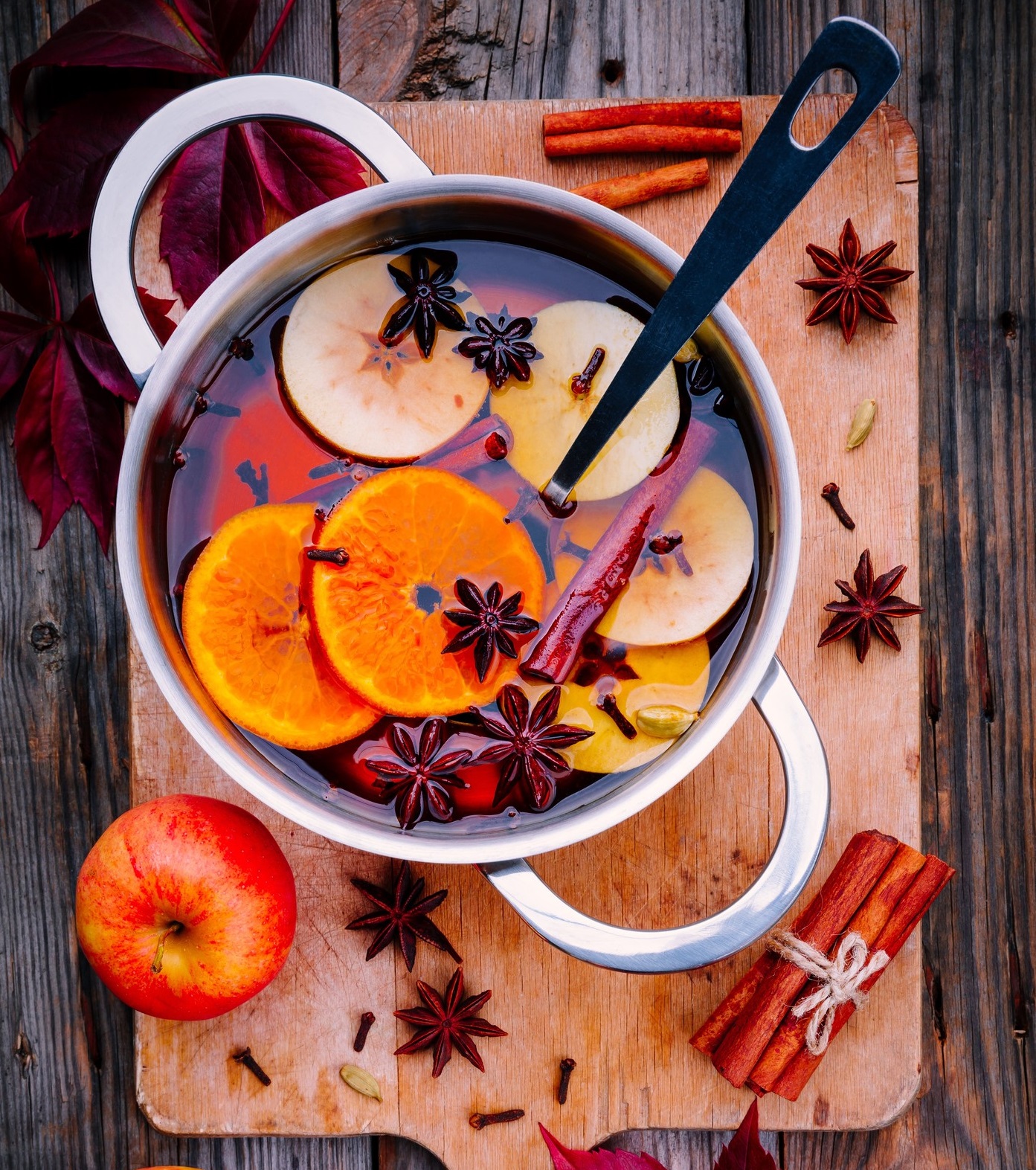 Hot mulled wine drink with citrus, apples, cinnamon sticks, cloves and anise in cooking pan on wooden background