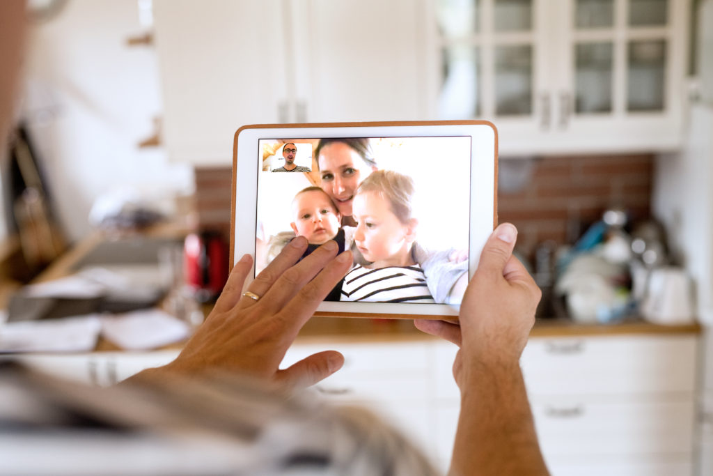 Unrecognizable young father at home holding a tablet, video chatting with wife and children.
