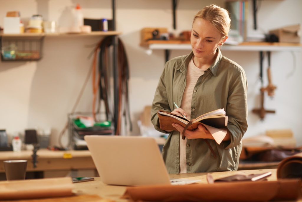 Horizontal shot of young female artisan looking at laptop screen writing something in her handmade notebook with leather cover