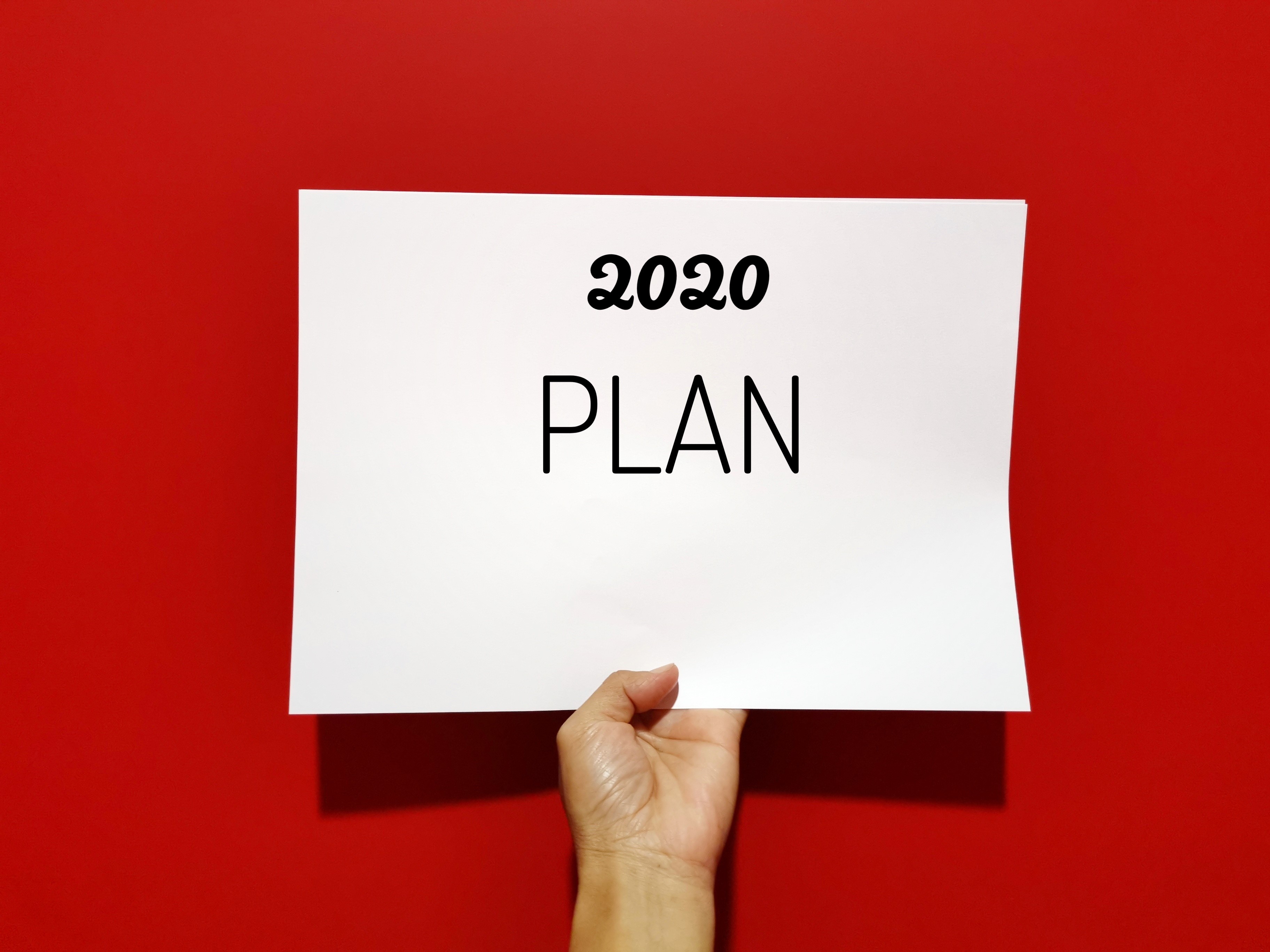 A hand holding up a white sign against a red background that says 2020 Plan in black.
