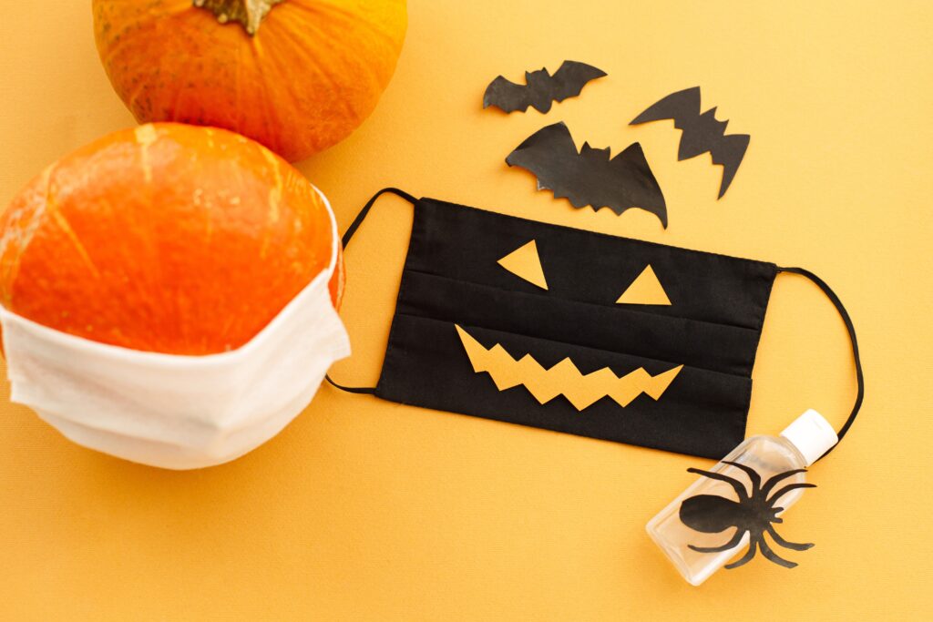 Halloween 2020 safe celebration. Evil face mask, pumpkin in mask, disinfection gel bottle, bat and spider decorations on orange background. Stay creative Stay safe