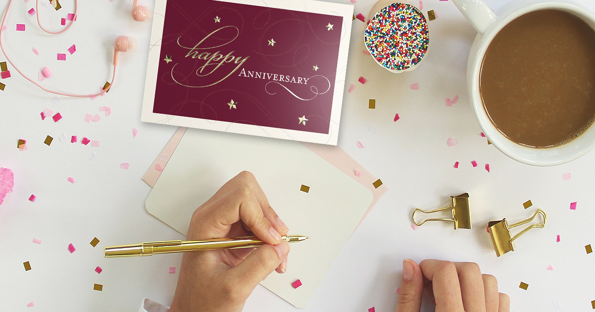 A hand holding a gold pen about to write a message in an anniversary card from CardsDirect.