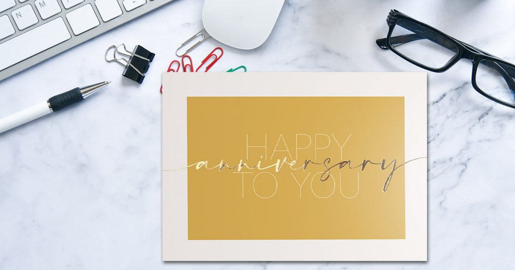 Gold anniversary card from CardsDirect on a marble background with colorful paperclips, reading glasses, a pen, and a computer.