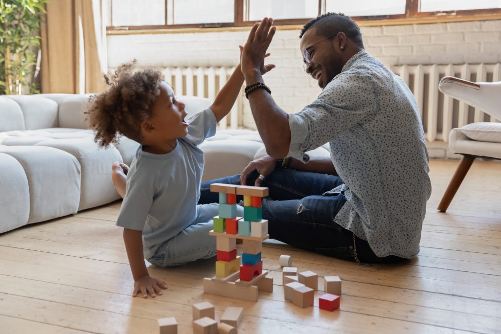 Full length playful little african american child boy giving high five to smiling young daddy, finishing constructing toy building with wooden bricks, sitting together on warm floor in living room.