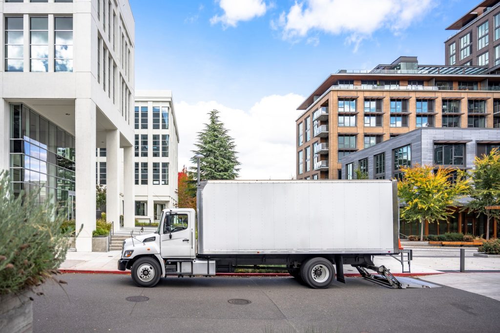 Relocation rig day cab semi truck tractor with long spacious box trailer standing on the urban city street with multilevel apartment and office buildings unloading delivered goods