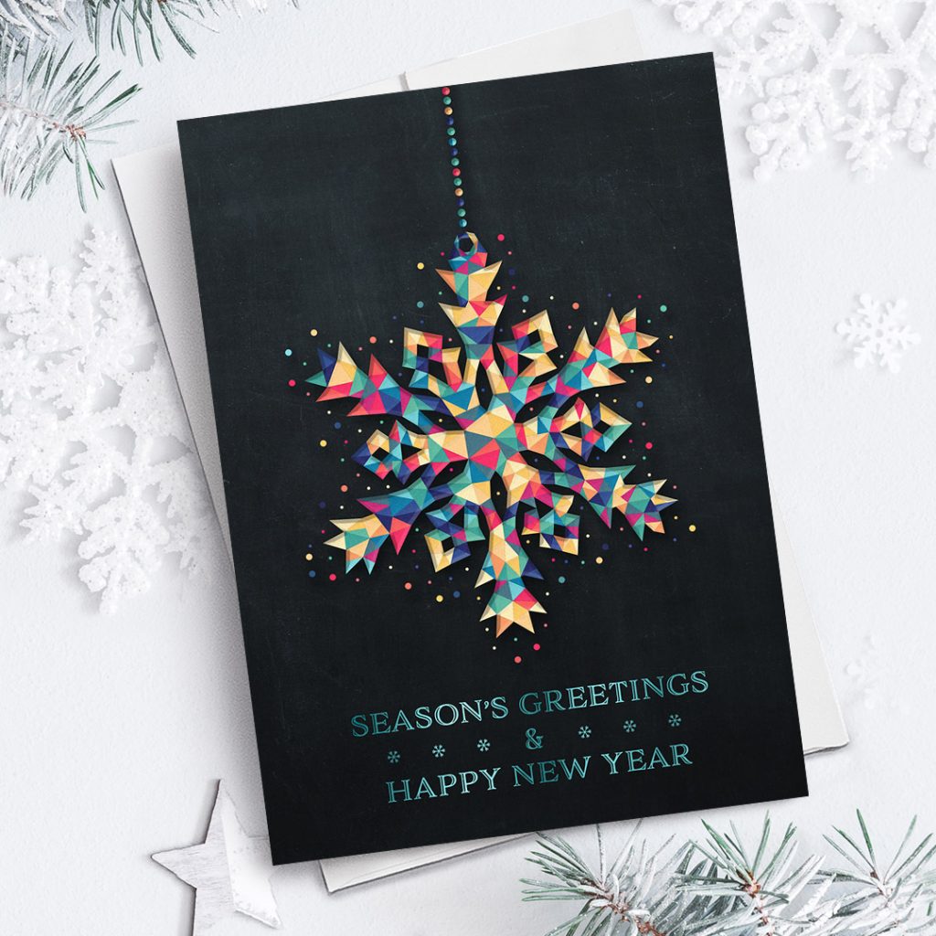 Christmas card with a colorful snowflake ornament on the front against a snow white background with pine sprigs, snowflakes, and stars