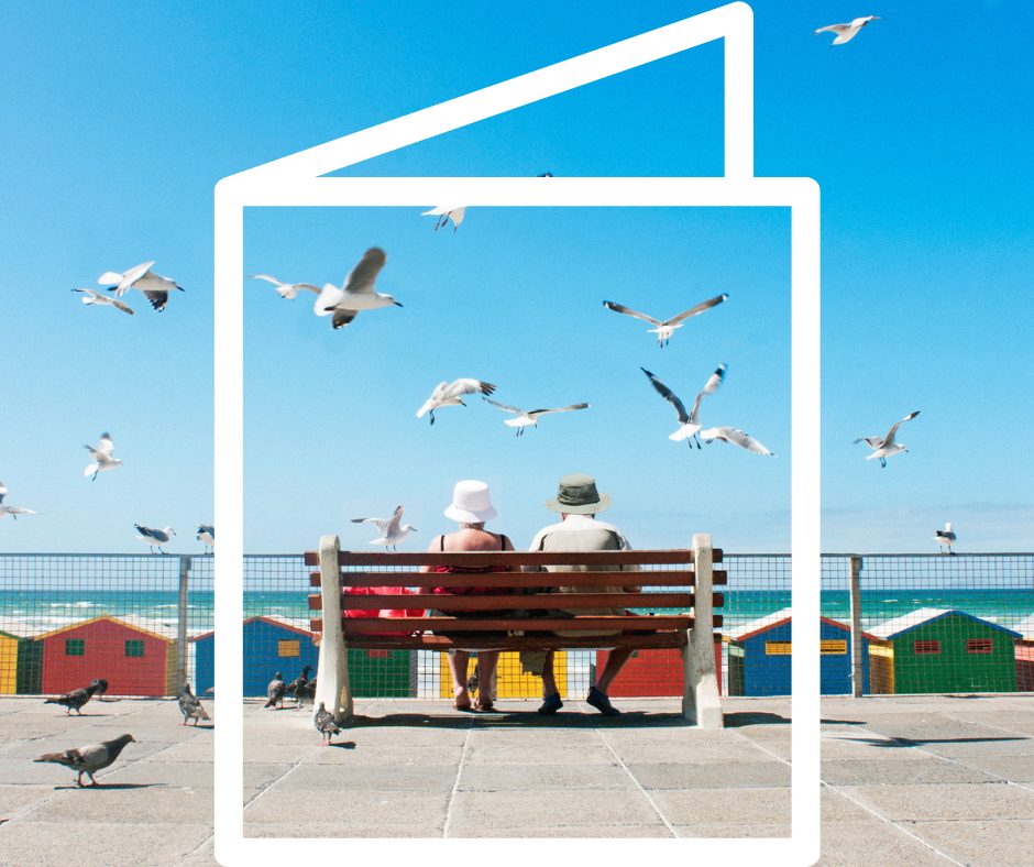 Elderly couple inside a greeting card frame sitting on a bench looking out over colorful houses on a beach with seagulls flying around.