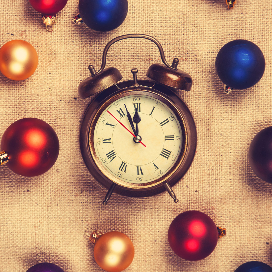 Antique clock about to strike 12 on a burlap background with colorful Christmas ornaments.